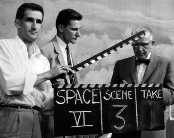 A historical production photograph of three men holding an open clapperboard marked scene three.